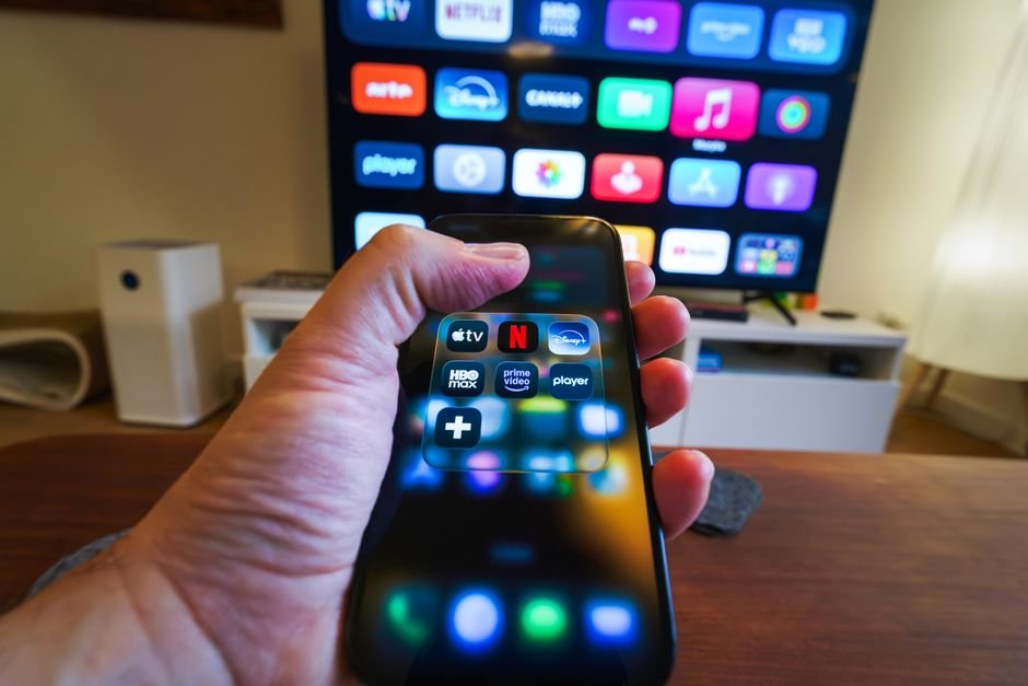 Hand holding a smartphone showing streaming app icons in front of a TV