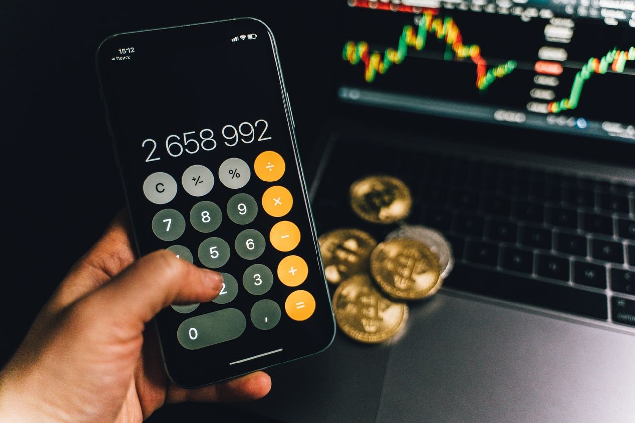 Calculator app beside bitcoin coins and a laptop chart, used for comparing IRA fee structures
